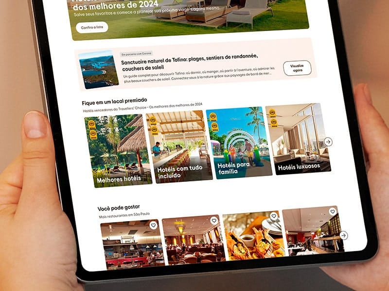 Pessoa segurando um tablet que exibe uma página de recomendações de hotéis no site TripAdvisor, incluindo categorias como melhores hotéis, hotéis com tudo incluído, hotéis para família e hotéis luxuosos.