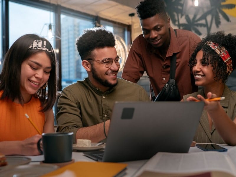 Quatro jovens adultos, três mulheres e um homem, colaborando alegremente em um projeto em frente a um laptop em um ambiente de trabalho descontraído, exemplificando princípios de trabalho em equipe e colaboração.