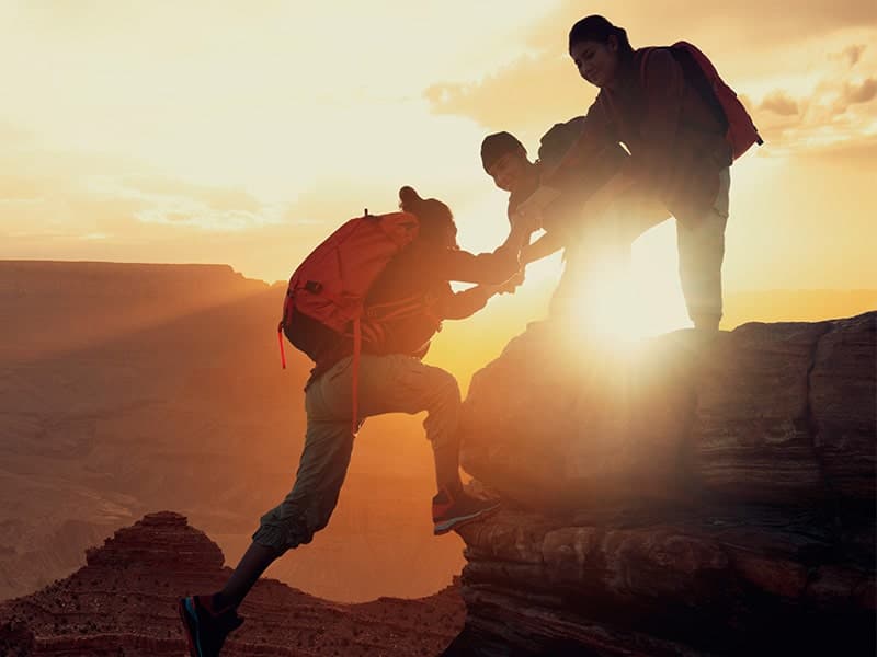 Três pessoas escalando uma montanha ao pôr do sol, demonstrando trabalho em equipe e liderança empresarial, com uma pessoa ajudando a outra a subir. As mochilas de trekking e a paisagem natural ao fundo enfatizam a superação de desafios e o espírito de colaboração.