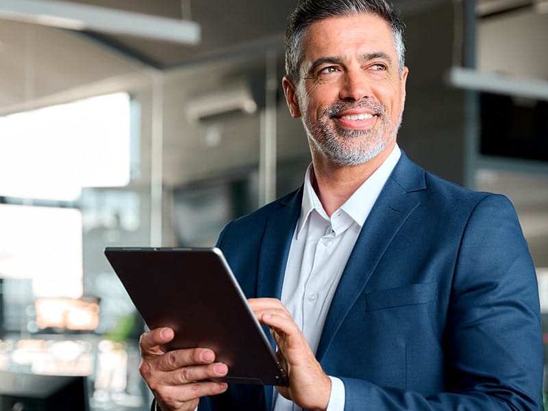 Homem de meia-idade, vestido com um terno azul, segurando um tablet e sorrindo enquanto olha para o lado. Ele está em um ambiente de escritório moderno e bem iluminado, transmitindo uma sensação de confiança e profissionalismo.