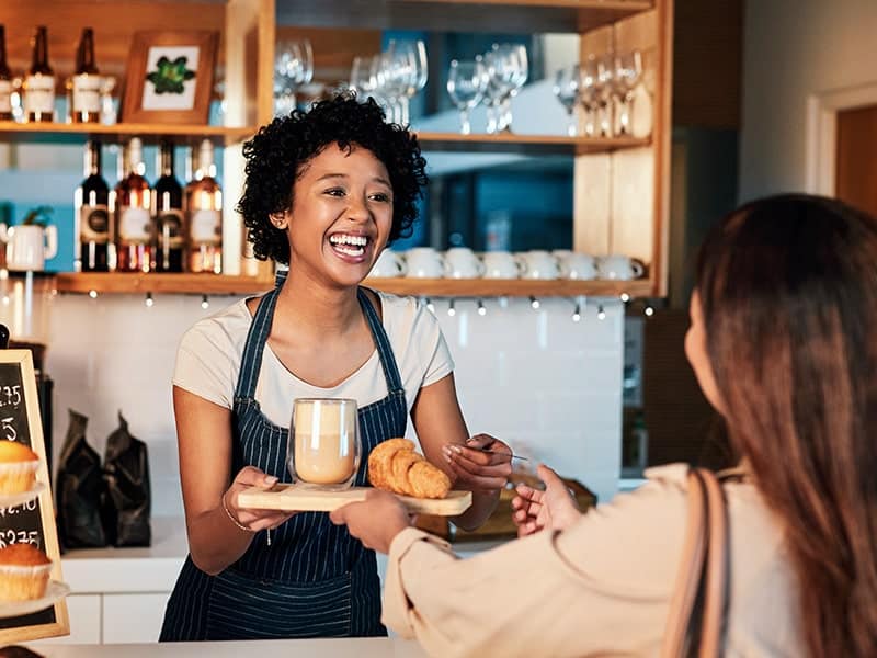 Uma barista sorridente entregando uma bandeja com um café e croissants para uma cliente em uma cafeteria acolhedora. A barista usa um avental listrado e está atrás do balcão, onde há prateleiras com garrafas de vinho e copos. A cliente, de costas para a câmera, está recebendo a bandeja com uma mão enquanto segura uma bolsa com a outra. A interação amigável entre as duas mulheres sugere um excelente atendimento ao cliente.