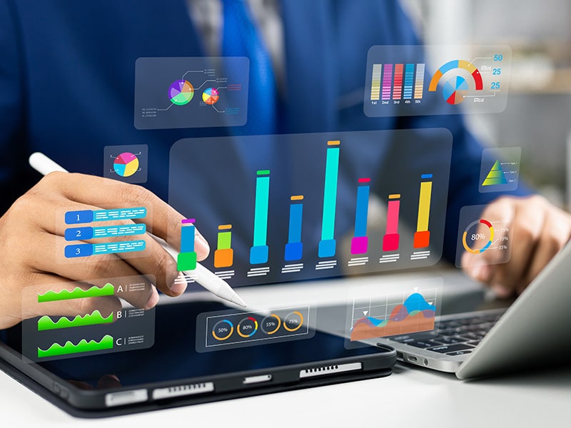 Pessoa utilizando um tablet para análise de gráficos e indicadores de desempenho, representando a aplicação da inteligência de dados em operações de negócios. Gráficos coloridos e dinâmicos flutuam acima do dispositivo, simbolizando o uso de tecnologia avançada para visualização e interpretação de grandes volumes de dados.